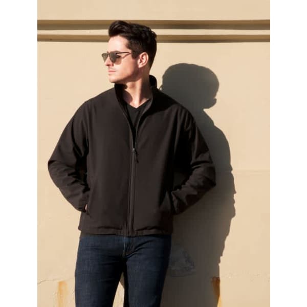 A man wearing a black soft shell jacket stands against a wall, with sunglasses and a shadow.