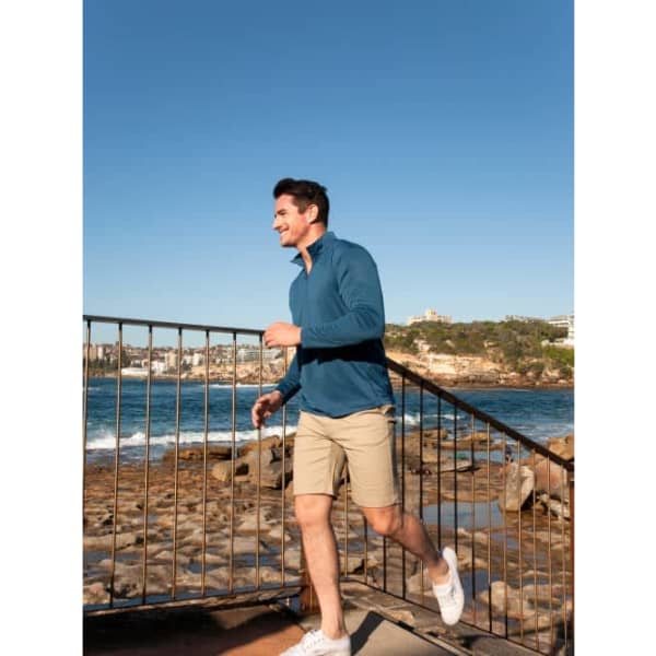 A man in a blue long-sleeve shirt and shorts walks near a rocky shoreline, with a waterfront city backdrop.
