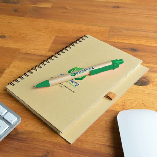 A brown spiral-bound notebook with a green pen placed on a wooden surface next to a keyboard and mouse.