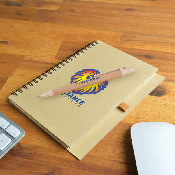 A brown spiral-bound notebook with a logo and a wooden pen resting on a wooden desk, next to a keyboard and mouse.