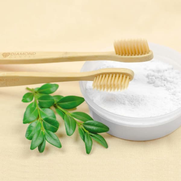 Two bamboo toothbrushes rest beside a bowl of white powder and green leaves on a beige surface.