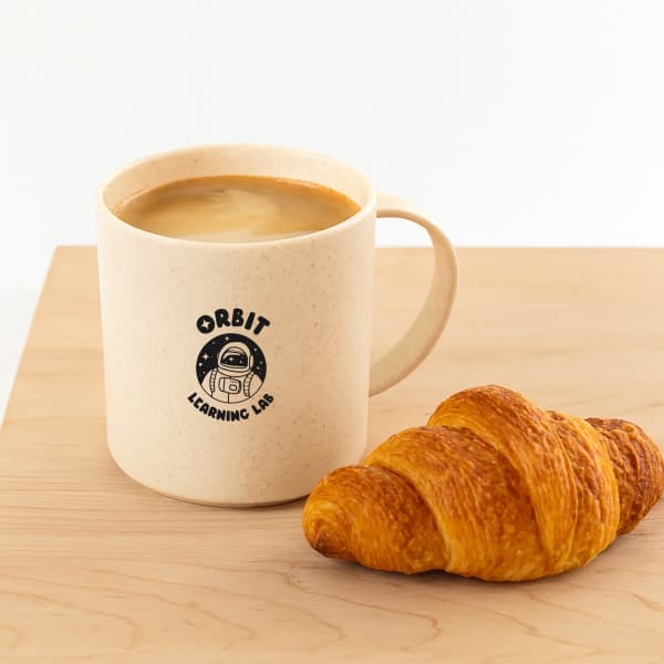 A cream-coloured reusable coffee mug with a logo, accompanied by a golden croissant on a wooden surface.
