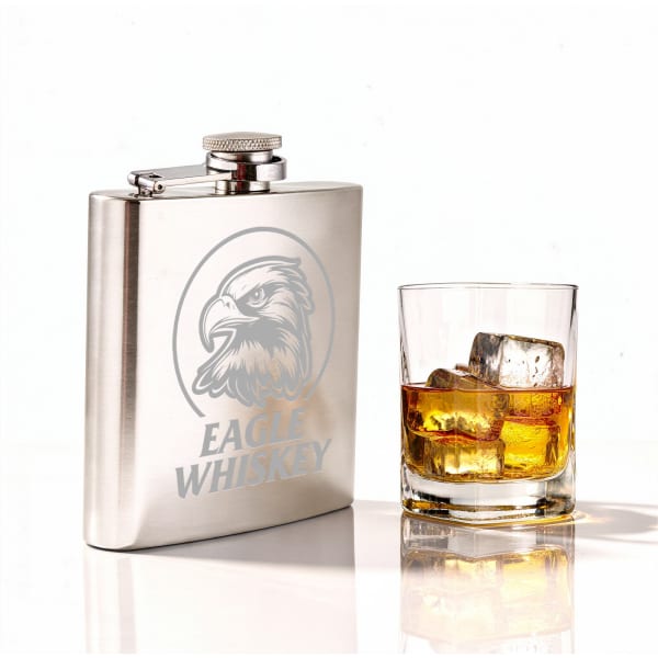 A stainless steel hip flask with an engraving, beside a glass of whiskey containing ice cubes.