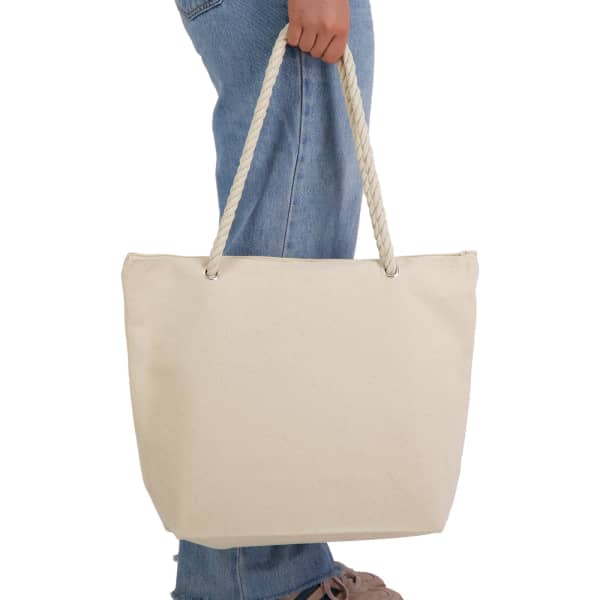 A cream-coloured canvas tote bag with rope handles, being held by a person wearing blue jeans.