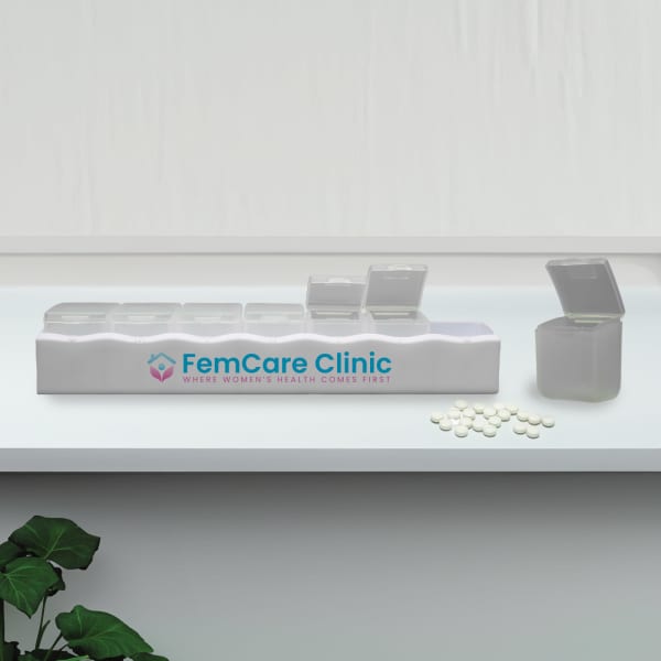 A white weekly pill box with multiple compartments, featuring a logo and a few pills scattered nearby.