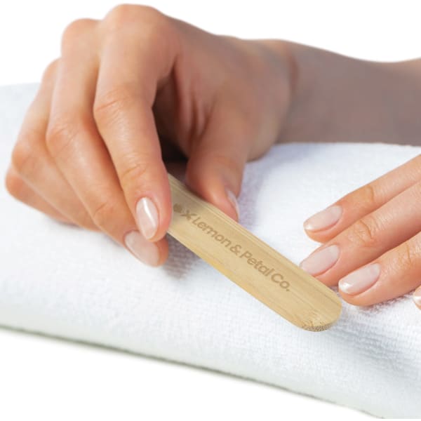A bamboo nail file held between fingers, resting on a white towel. The file has a logo on its side.
