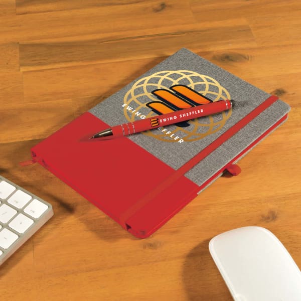 A grey notebook with a red elastic band and a matching red pen placed on a wooden desk beside a keyboard and mouse.