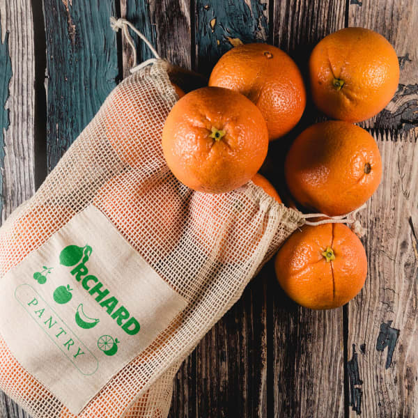A mesh produce bag in natural colour, filled with bright orange oranges, displayed on a rustic wooden surface.