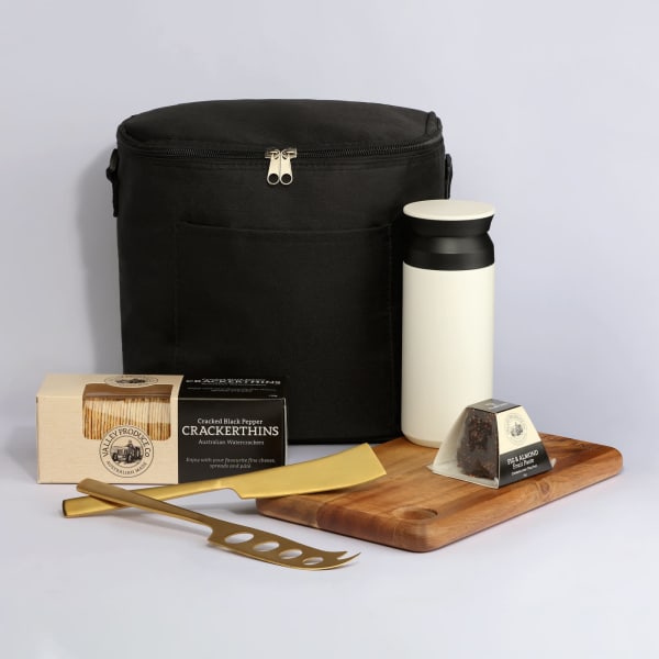 A black cooler bag with a drink bottle, wooden serving board, cheese, crackers, and a gold cheese knife.