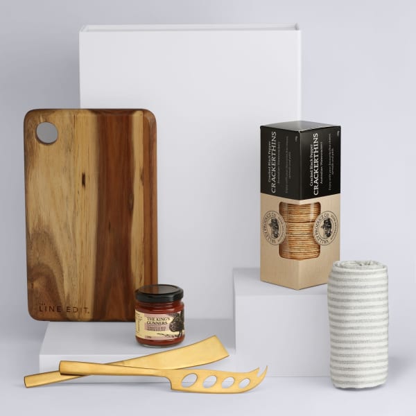 A wooden cheese board, two cheese knives, a jar of chutney, and a box of crackers, presented on white pedestals.