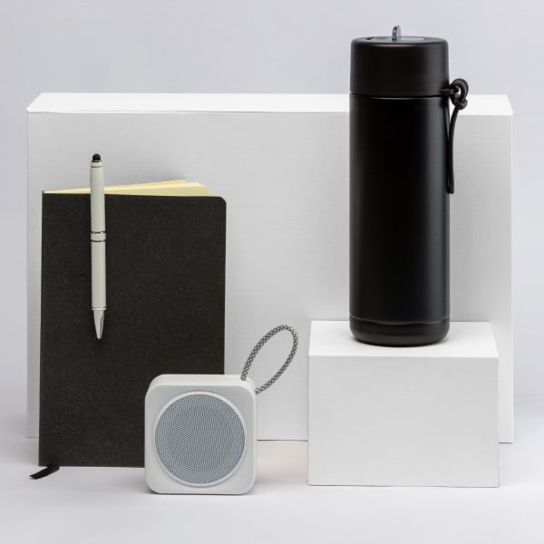 A black drink bottle, a black notebook, a silver pen, and a white speaker arranged on white blocks.