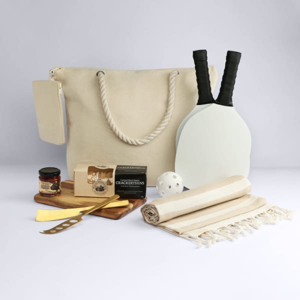 A cream tote bag with rope handles, two paddles, a ball, serving board, knife, and picnic blanket.
