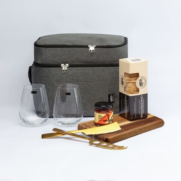 A grey cooler bag with two glasses, a wooden serving board, cheese, crackers, and a jar. The bag features a logo.