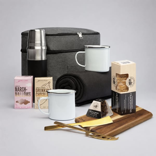 A black cooler bag with a drink bottle, mugs, marshmallows, chocolate drink mix, a cheese board, and utensils.