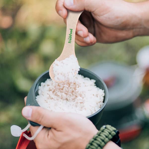 A person holds a beige wheat fibre spoon above a bowl of uncooked rice. The utensil has a logo.