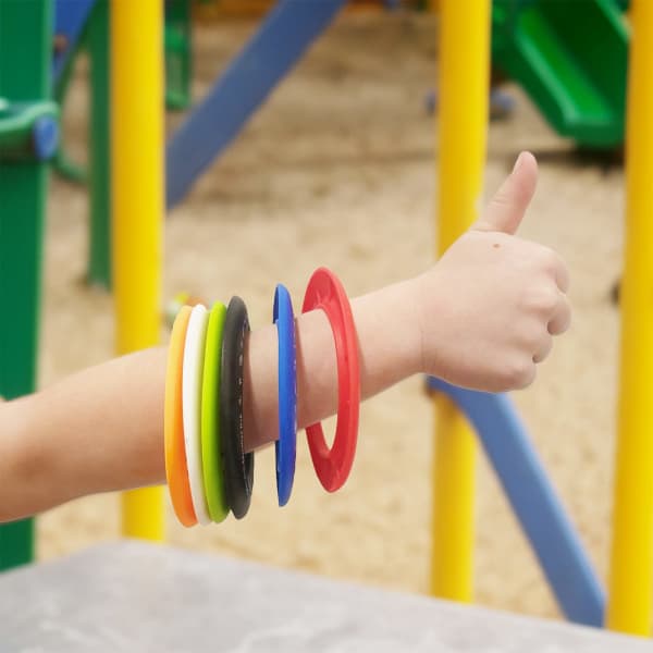 A wrist adorned with colourful silicone flying discs in red, green, yellow, black, and blue, set against a playground backdrop.