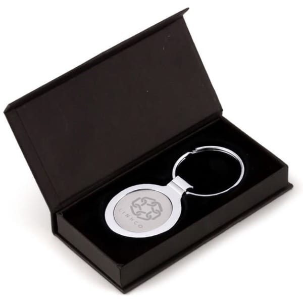 A round silver keyring displayed in a black gift box, featuring a logo on the keyring.