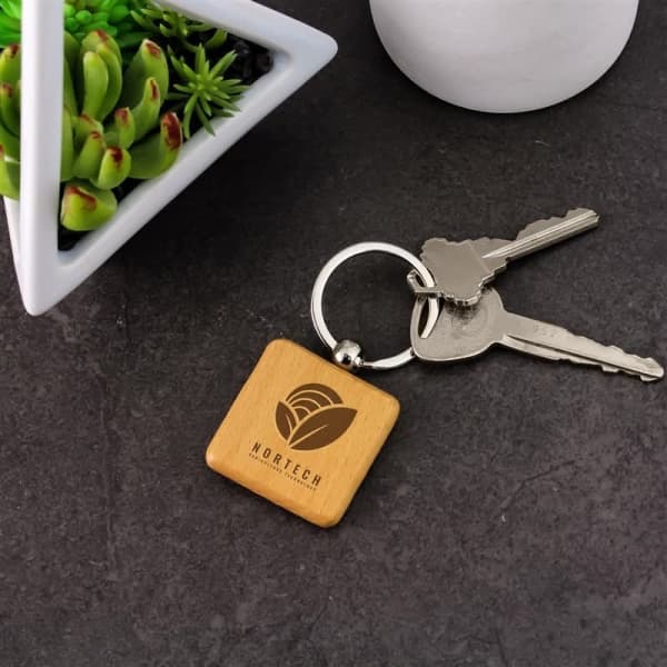 A square wooden keyring with a metal ring, accompanied by silver keys, and a small green plant in the background.