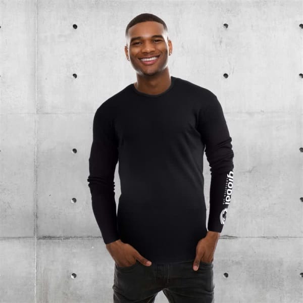 A long-sleeve black shirt with a logo on the sleeve, worn by a smiling person against a concrete backdrop.