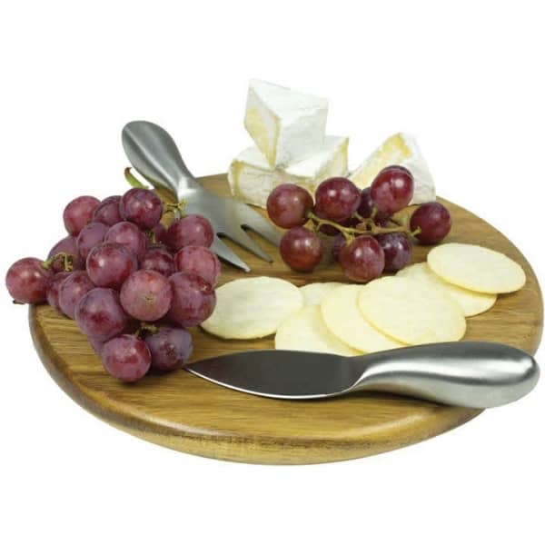 A wooden round cheese board featuring slices of cheese, grapes, and cheese knives.