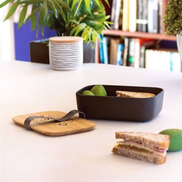 A black bamboo fibre lunch box with a wooden lid, containing a sandwich and green apples, on a table with plants.