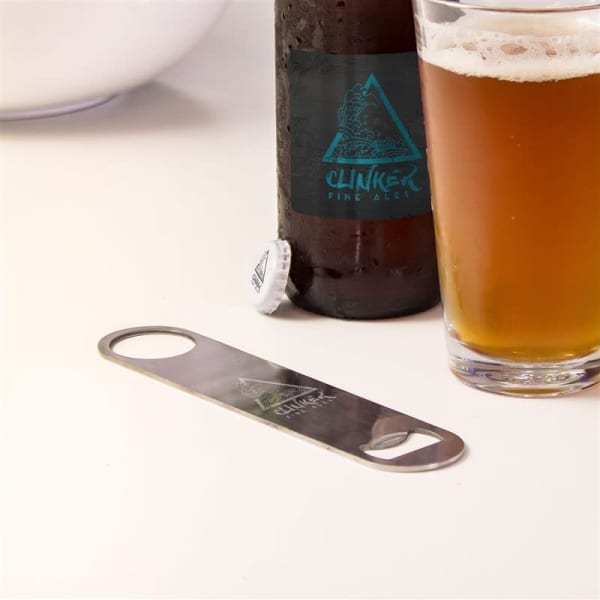 A drink bottle opener in silver beside a brown beer bottle and a half-filled glass of amber beer on a white table.