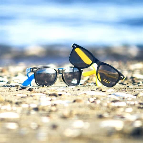 Retro sunglasses in black and blue, resting on sandy beach with a blurred ocean background.