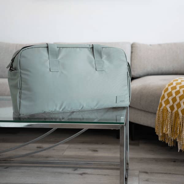 A green duffle bag sits on a glass table, with a sofa and blanket in the background. The bag has handles and a logo.