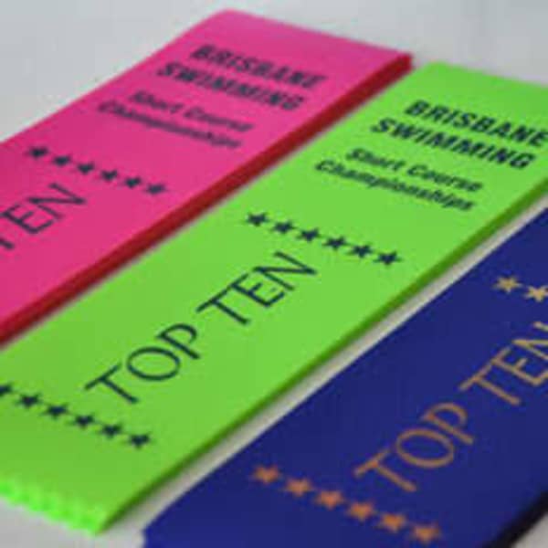 Three 15cm long award ribbons in bright pink, neon green, and blue, each featuring printed text and a logo.