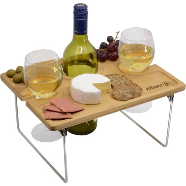 A wooden picnic table with two glasses of white wine, a bottle, cheese, crackers, olives, and grapes.