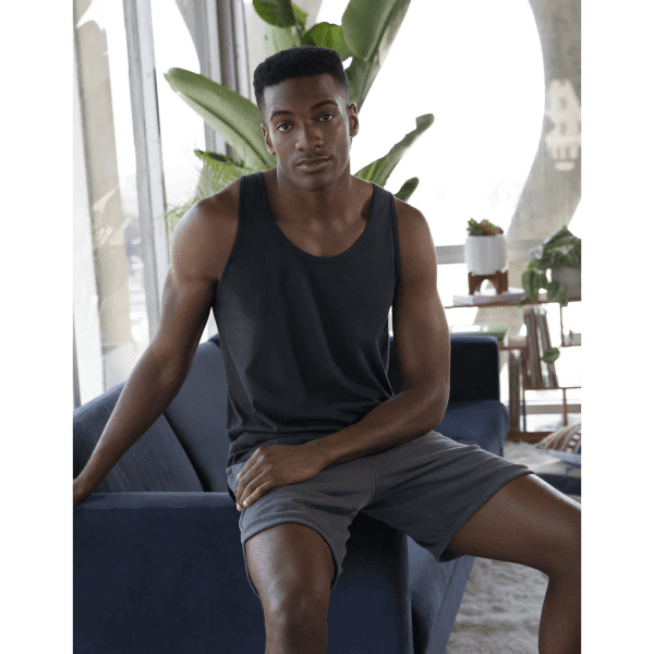 A black unisex tank top is worn by a model sitting on a blue couch, surrounded by indoor plants.