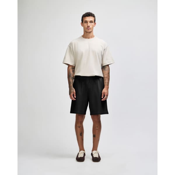A man stands wearing a beige t-shirt and black gym shorts, showcasing a simple, casual outfit. The shorts feature a logo.