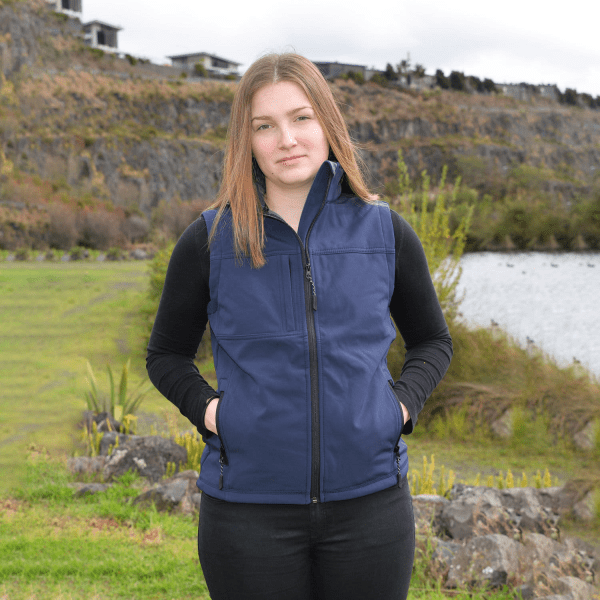 A navy blue softshell vest with a zip and pockets, worn by a model in an outdoor setting near water and greenery.