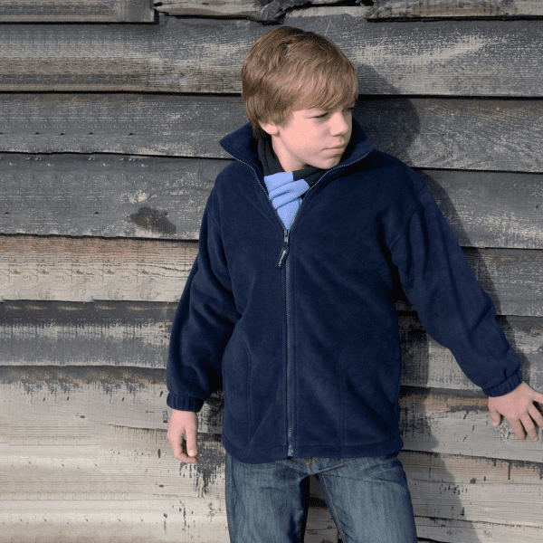 A youth wearing a navy blue polar fleece full zip jumper, standing against a wooden wall. The jumper has a logo.