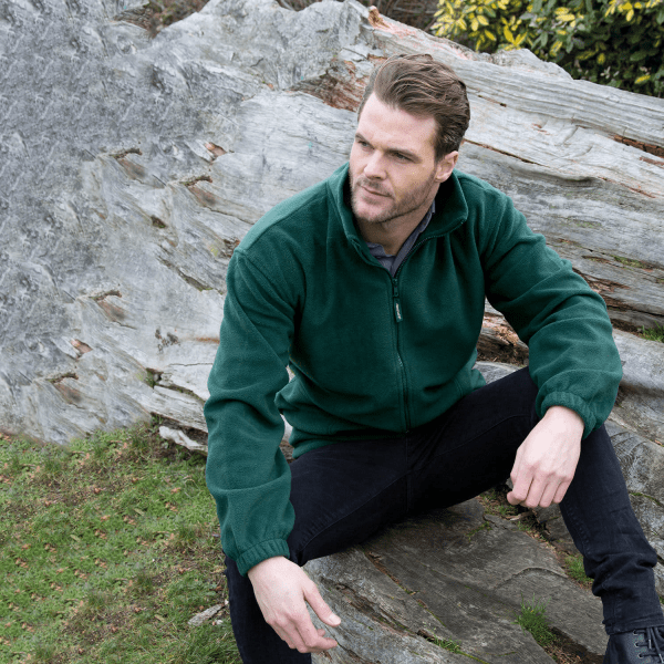 A green polar fleece full zip jumper is displayed on a model sitting on a rock in a natural setting.