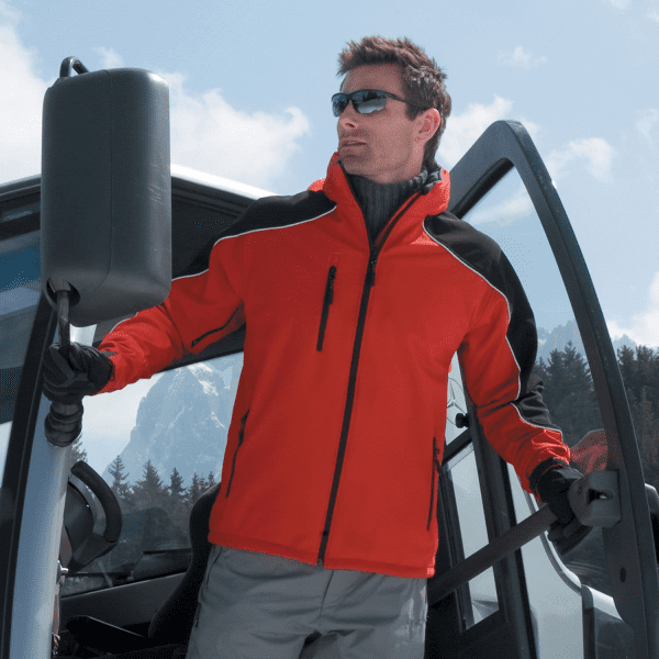 A man wearing a red softshell jacket with black accents stands by a vehicle in a mountainous setting.