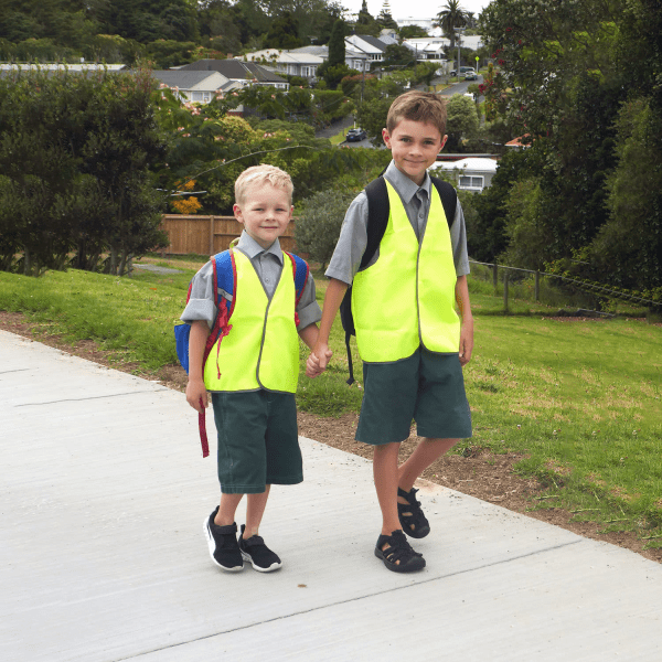 Two children wearing bright yellow safety vests and school uniforms, walking hand in hand on a path.