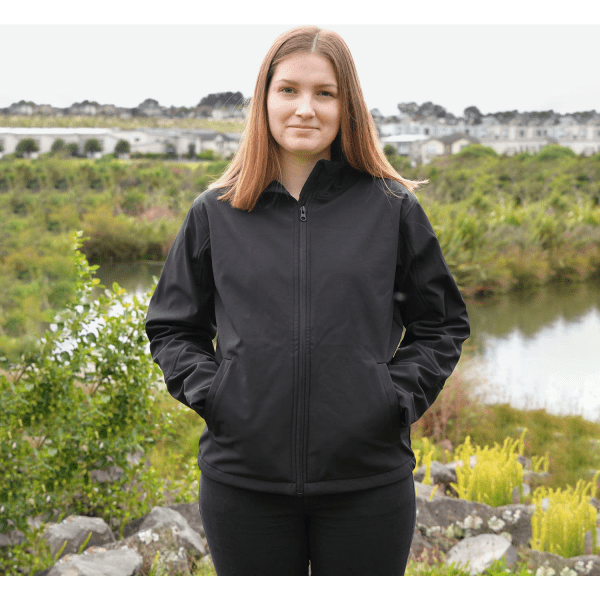 A black softshell jacket is worn by a woman standing outdoors, surrounded by greenery and water. The jacket has a logo.