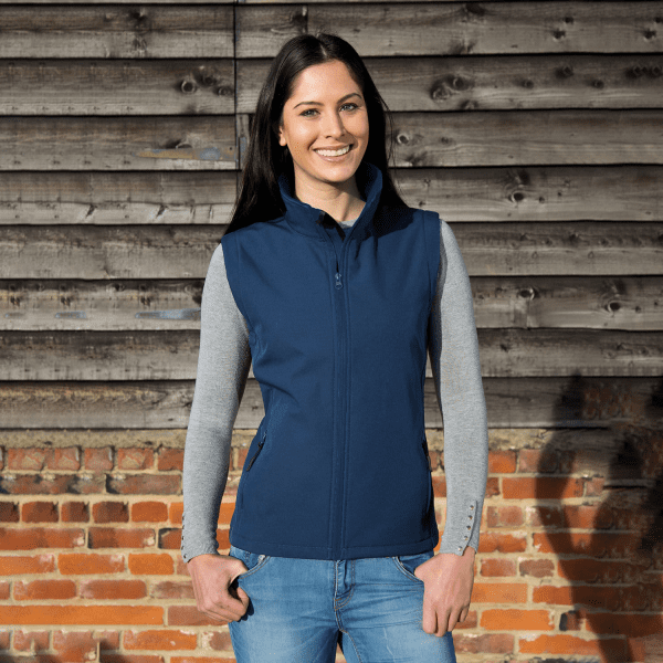 A ladies' softshell vest in navy blue, featuring a zip front and a fitted design, displayed against a rustic wooden backdrop.
