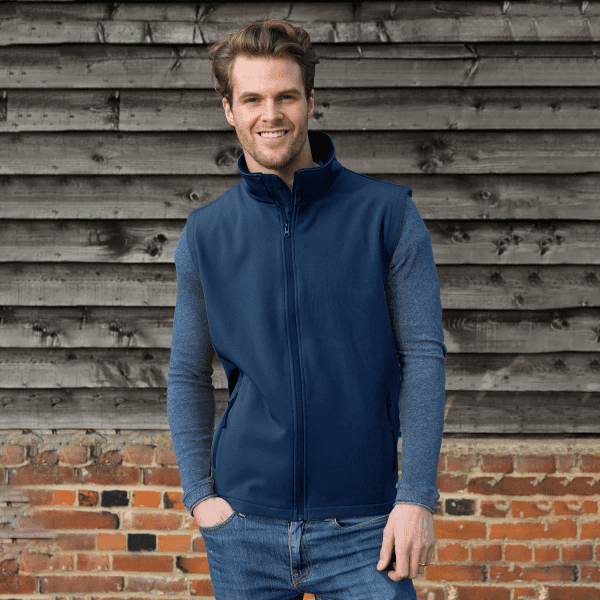 A navy softshell vest features a high collar and zip closure, paired with long grey sleeves. It has a logo on the chest.