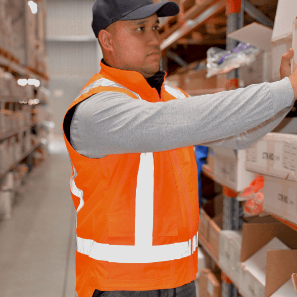 A menswear model wearing an orange reversible fleece lined safety vest with reflective stripes in a warehouse setting.