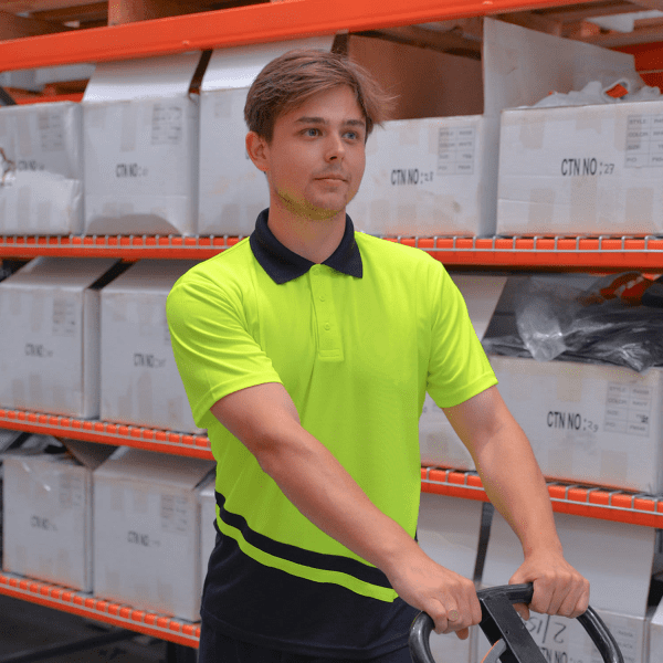A man in a high-visibility yellow and navy polo shirt stands next to a trolley in a warehouse. The shirt has a logo.