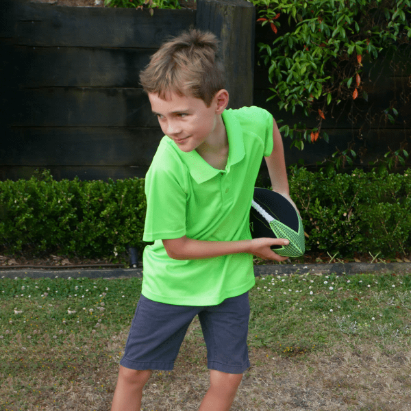 A young boy wearing a bright green polo shirt and grey shorts is holding a rugby ball while posing outdoors.