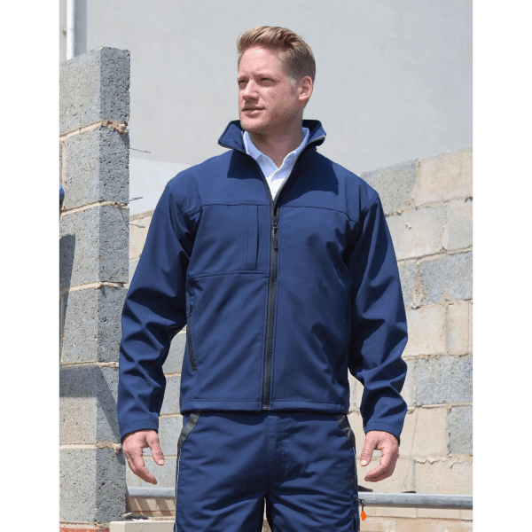 A man in a navy soft shell jacket stands outdoors near a stone wall, featuring a logo on the jacket.
