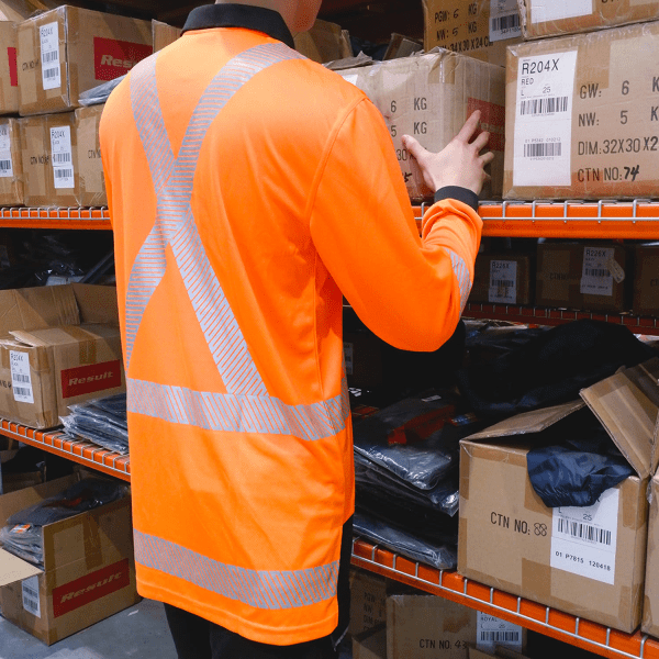 A person wearing an orange long sleeve polo shirt with reflective stripes, standing near cardboard boxes on a shelf.