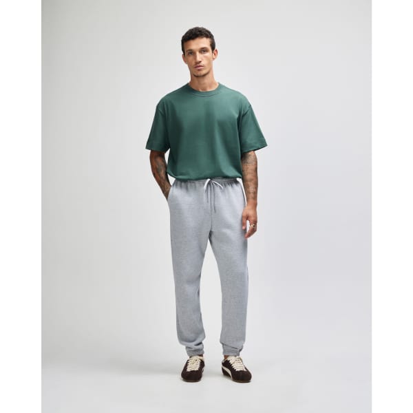 A man stands wearing grey unisex sweatpants and a green t-shirt, showcasing a casual outfit. The sweatpants have a logo.