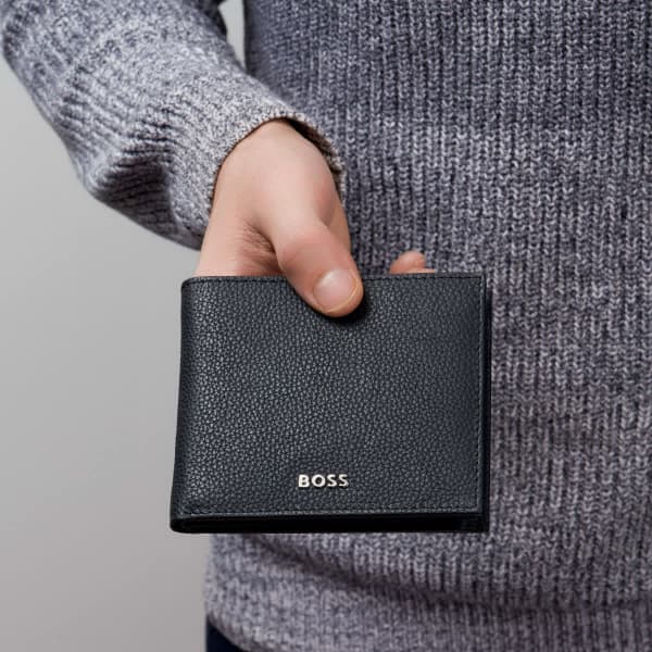 A black grained leather wallet held by a person wearing a grey sweater, featuring a subtle logo.