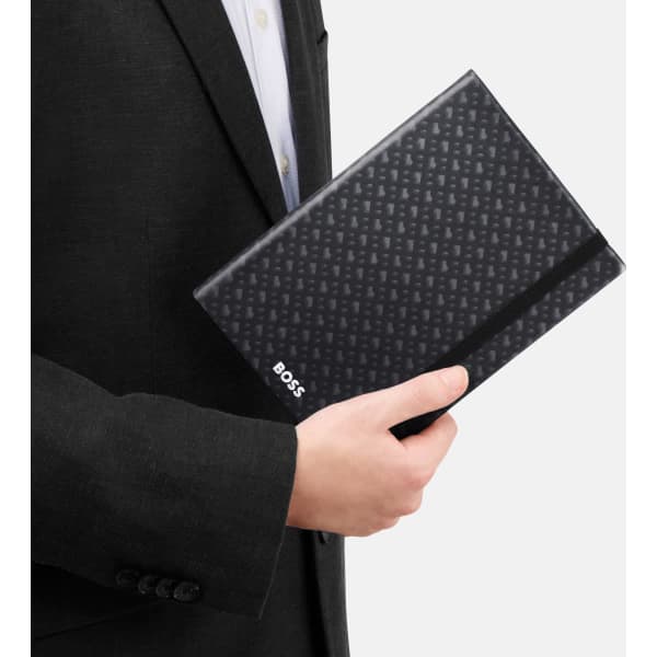 A black A5 notepad with a monogram pattern, held by a person in a suit. The notepad features a branding logo.