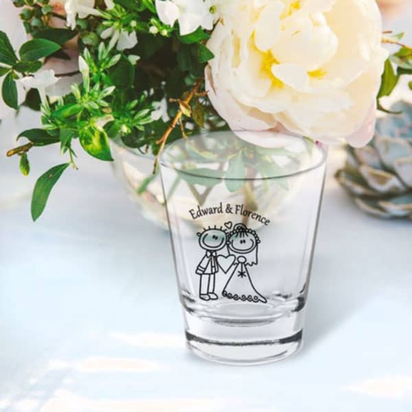 A clear shot glass featuring a couple illustration, surrounded by flowers. The glass has a logo.