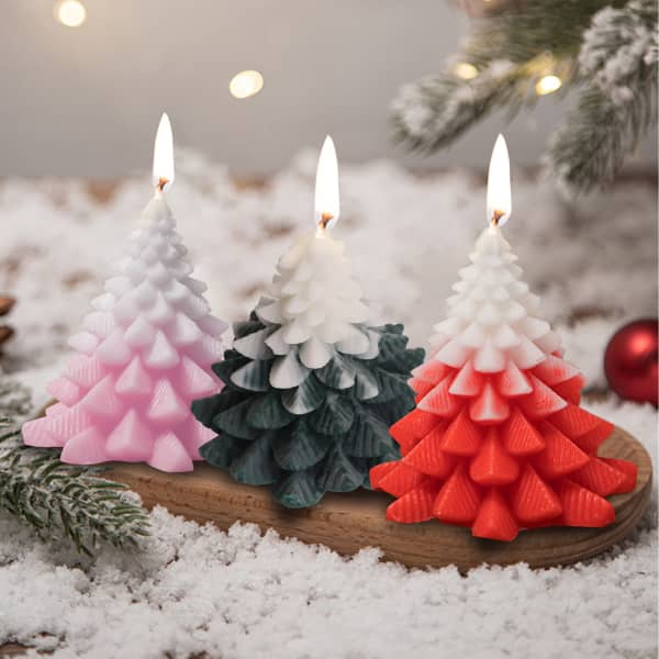 Three Christmas tree shape candles in pink, green, and red, with flames, displayed on a wooden board among snow and decorations.